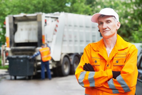 A male waste management worker stands outdoors in the foreground, wearing an orange high-visibility jacket with reflective silver and orange stripes, black gloves, and a white cap. His arms are crossed as he faces the camera with a neutral expression. In the background, there is a large white rubbish collection truck with its rear loading compartment open, indicating active rubbish collection, and another worker dressed in high-visibility clothing is seen near the truck. The setting appears to be a paved driveway or street with green foliage and trees visible beyond, suggesting a suburban or urban environment, possibly within the Spitalfields area. Natural daylight illuminates the scene, highlighting the textures of the high-visibility clothing, the metallic surface of the rubbish truck, and the surrounding greenery. The image underscores professional waste collection activities consistent with rubbish removal services offered by companies like Office Clearance Spitalfields.