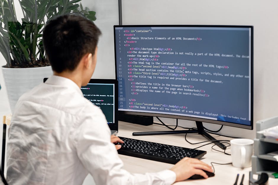 The image depicts a person with short dark hair, dressed in a white shirt, working at a desk in an office environment. They are sitting in front of a dual-monitor setup, with one large screen displayi