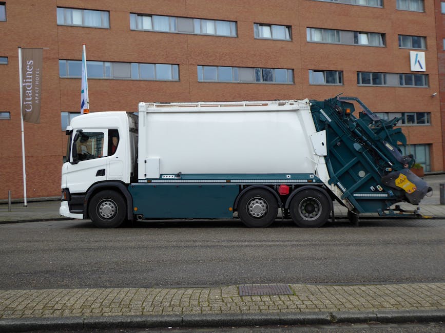 A street scene showing a waste collection truck with a large, open rear compartment that appears to be used for the collection of mixed rubbish. The truck is predominantly white with some patches of r